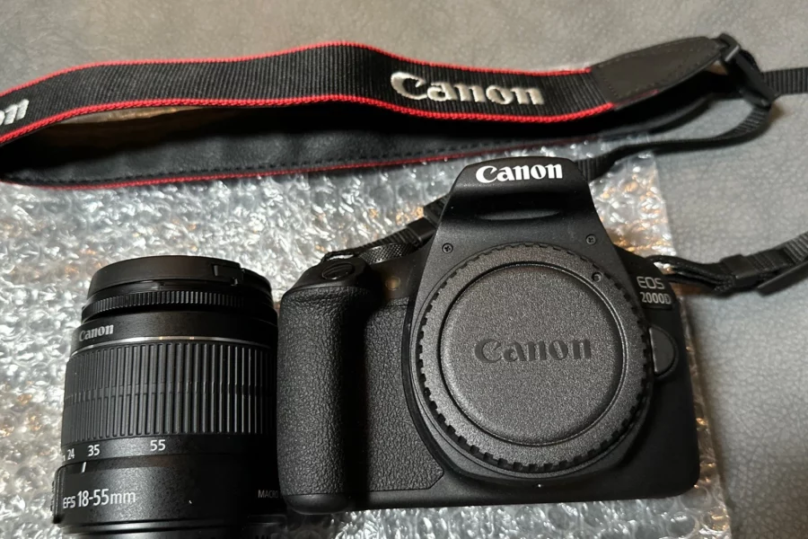 A flat lay shot of a Canon EOS 2000D camera kit on a gray surface with bubble wrap. The kit includes a black Canon EOS 2000D camera body with a lens cap on, a separate Canon EF-S 18-55mm lens, and a black and red Canon camera strap.
