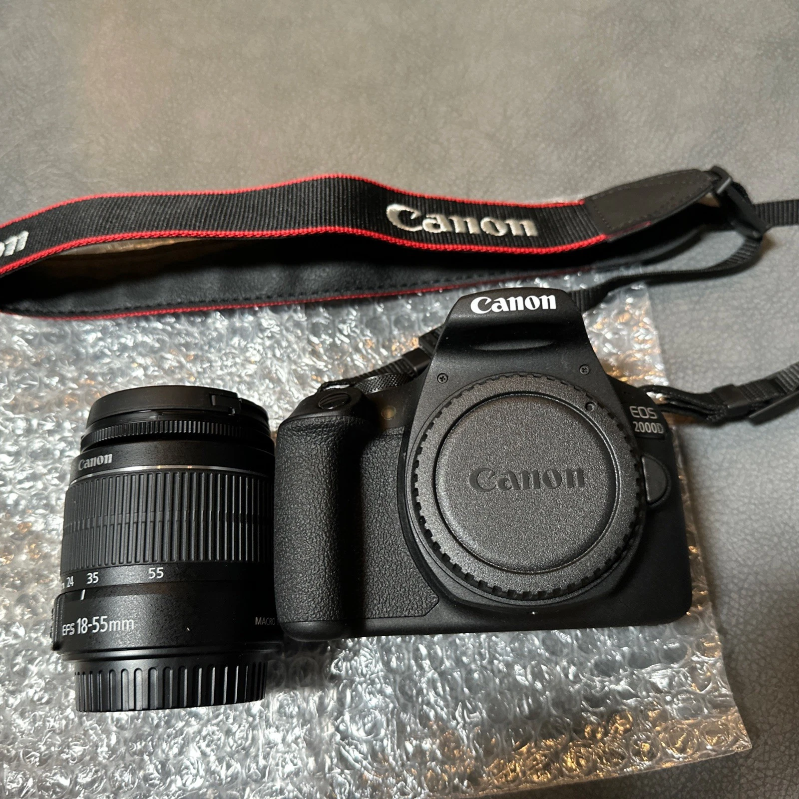 A flat lay shot of a Canon EOS 2000D camera kit on a gray surface with bubble wrap. The kit includes a black Canon EOS 2000D camera body with a lens cap on, a separate Canon EF-S 18-55mm lens, and a black and red Canon camera strap.