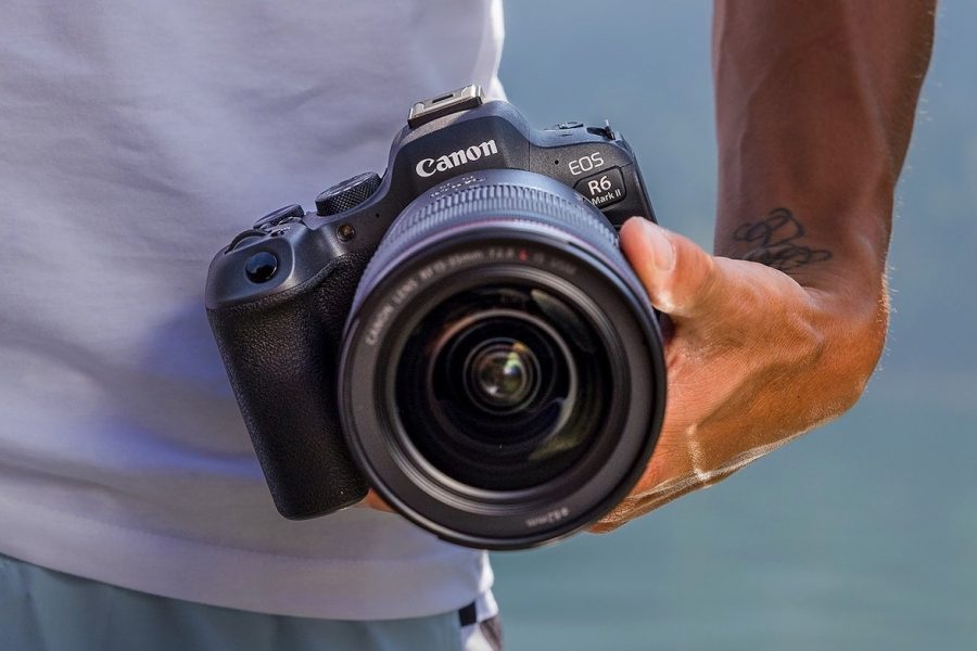 A low-angle close-up shot of a person's hand holding a Canon EOS R6 Mark II camera. The lens is facing the viewer and the person's tattooed forearm is visible in the frame. The person is wearing a white T-shirt.