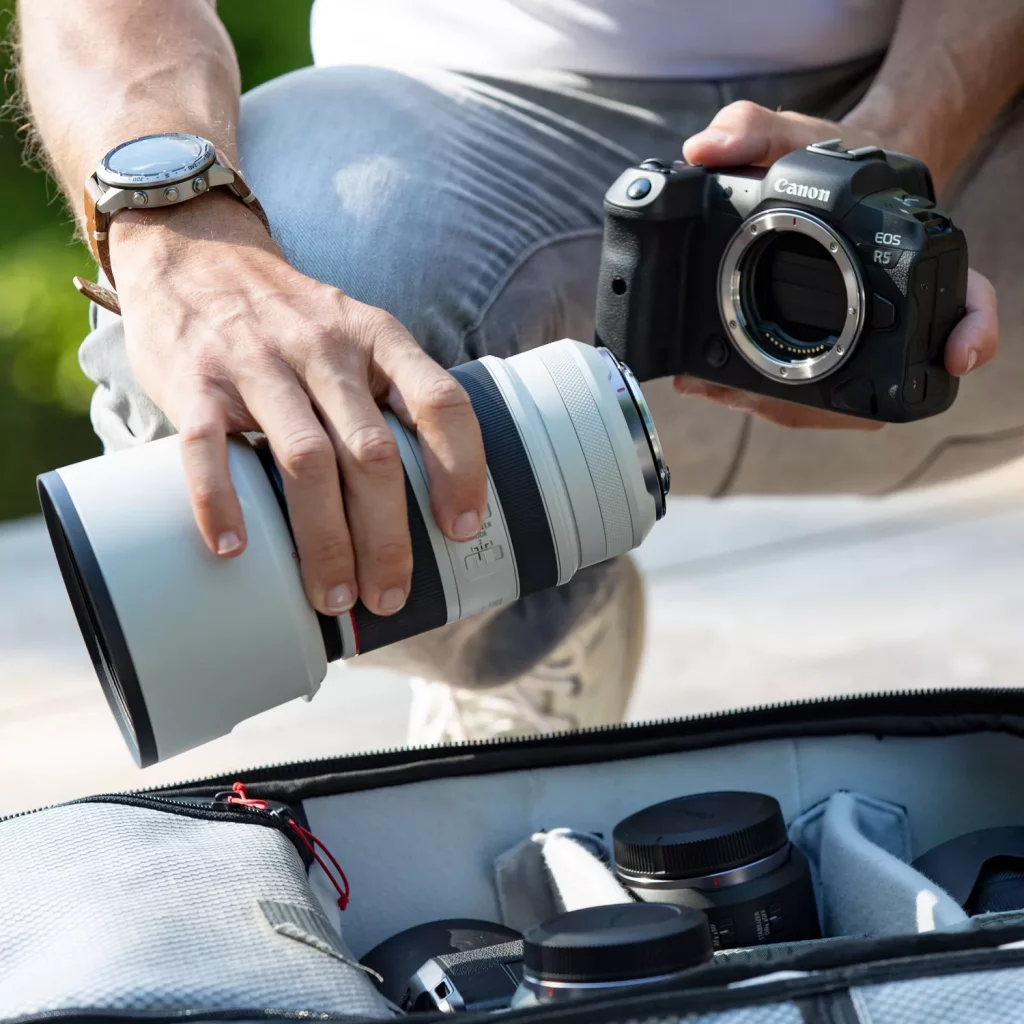 A photographer, wearing a white t-shirt, gray pants, and a wristwatch, is in the process of taking a white Canon RF 70-200mm F2.8 L IS USM lens out of a gray camera bag. He holds a black Canon EOS R5 camera body in his other hand. Other lenses and camera accessories are visible inside the bag.