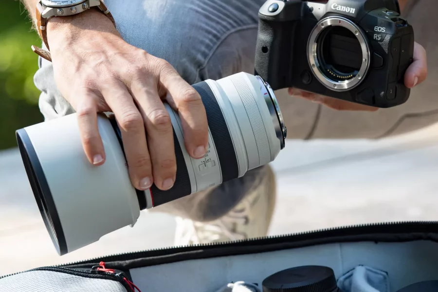 A photographer, wearing a white t-shirt, gray pants, and a wristwatch, is in the process of taking a white Canon RF 70-200mm F2.8 L IS USM lens out of a gray camera bag. He holds a black Canon EOS R5 camera body in his other hand. Other lenses and camera accessories are visible inside the bag.