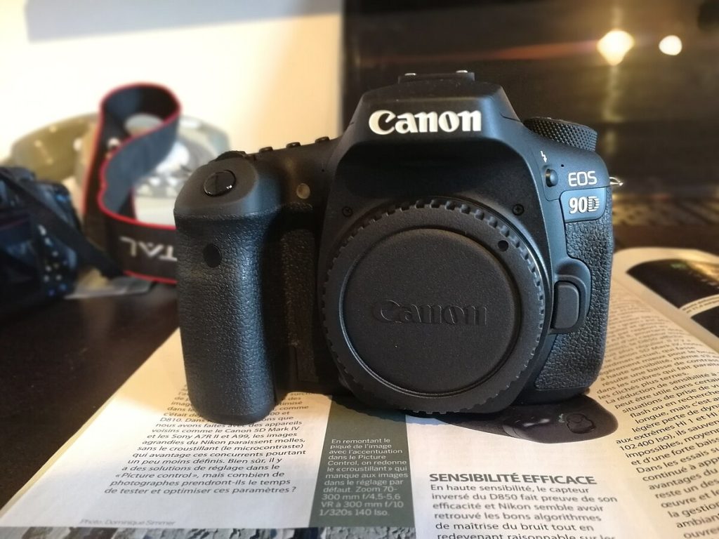 A close-up studio shot of a Canon EOS 90D camera body, positioned at a slight angle against a white background. The camera is shown without a lens, and a black body cap is attached.