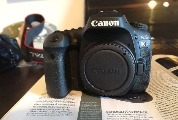 A close-up studio shot of a Canon EOS 90D camera body, positioned at a slight angle against a white background. The camera is shown without a lens, and a black body cap is attached.