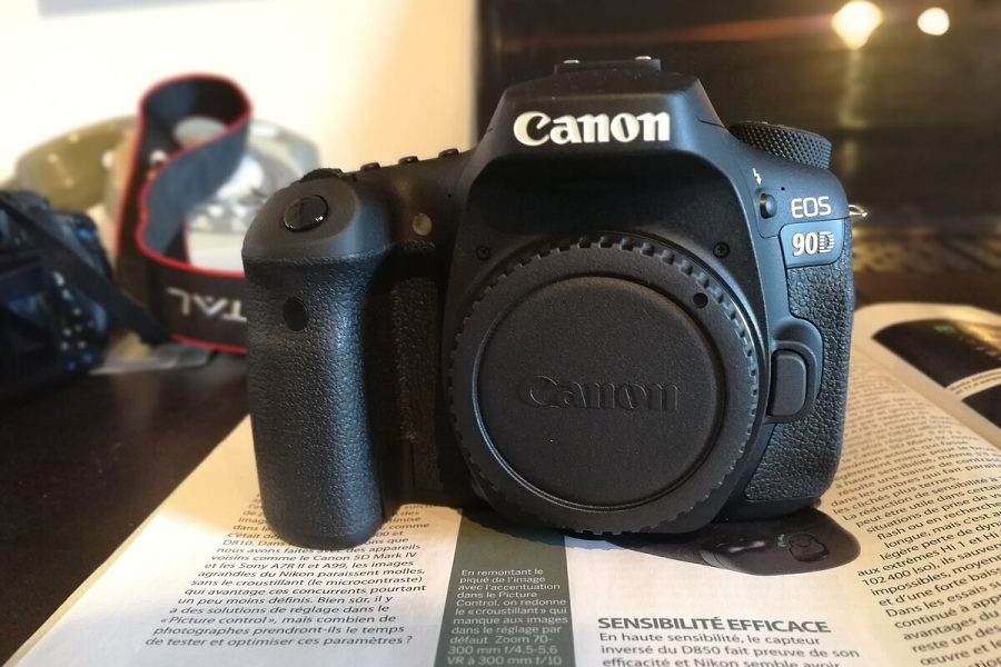 A close-up studio shot of a Canon EOS 90D camera body, positioned at a slight angle against a white background. The camera is shown without a lens, and a black body cap is attached.
