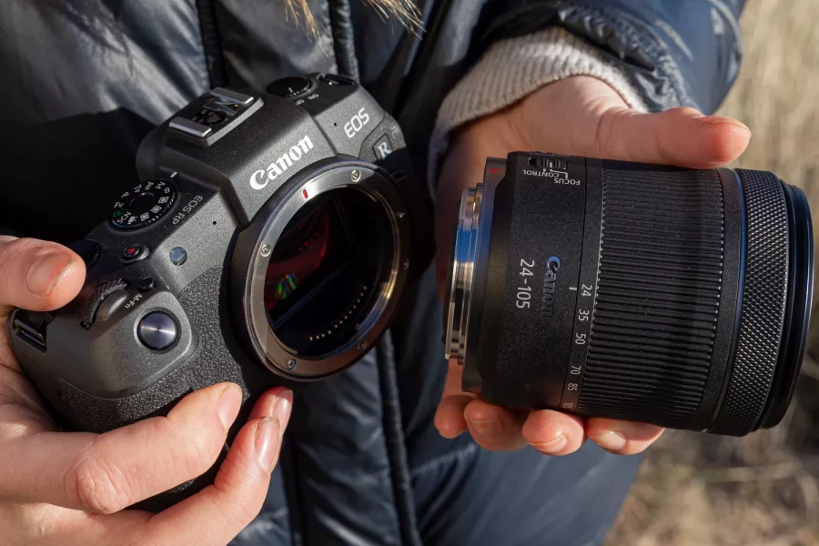 A pair of hands holds a Canon EOS RP camera body and an RF 24-105mm lens, ready to attach the lens to the camera. The camera's sensor is visible within the open lens mount. The person holding the camera is wearing a black puffer jacket, and the background is a grassy field.
