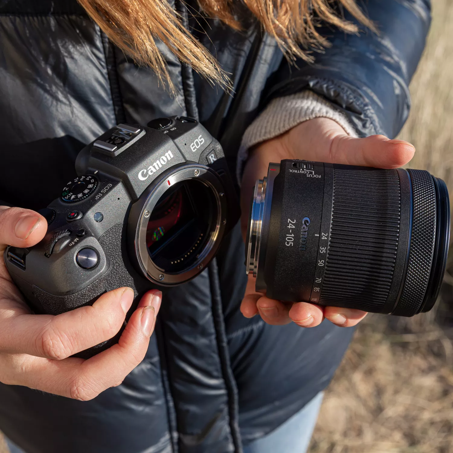 A pair of hands holds a Canon EOS RP camera body and an RF 24-105mm lens, ready to attach the lens to the camera. The camera's sensor is visible within the open lens mount. The person holding the camera is wearing a black puffer jacket, and the background is a grassy field.