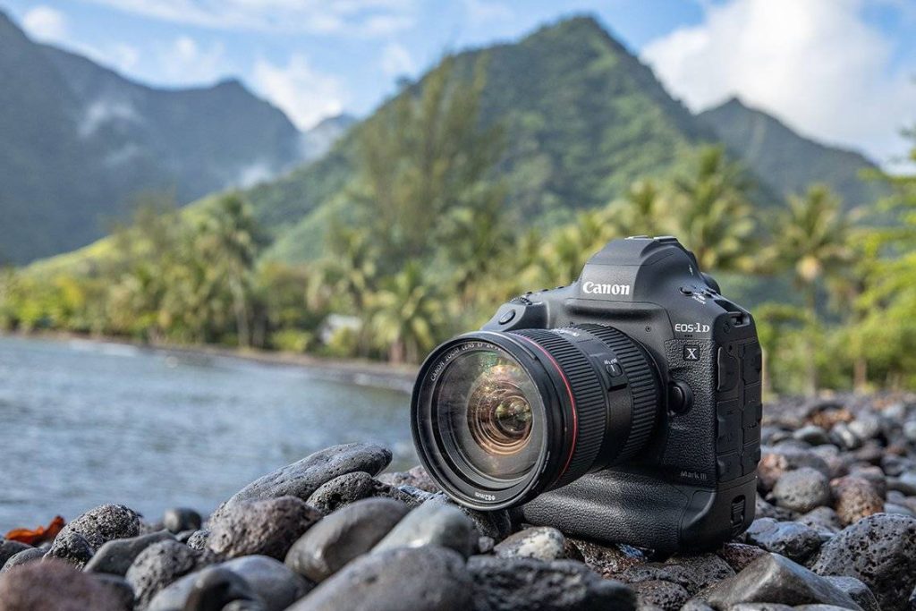 A close-up shot of a black Canon EOS-1D X Mark III DSLR camera with a lens attached, resting on a rocky beach. In the background, there's a body of water (likely the ocean or a lake) and lush green mountains under a partly cloudy sky. Palm trees are visible along the shoreline.