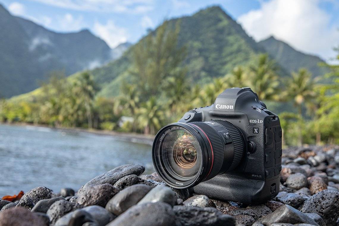 A close-up shot of a black Canon EOS-1D X Mark III DSLR camera with a lens attached, resting on a rocky beach. In the background, there's a body of water (likely the ocean or a lake) and lush green mountains under a partly cloudy sky. Palm trees are visible along the shoreline.