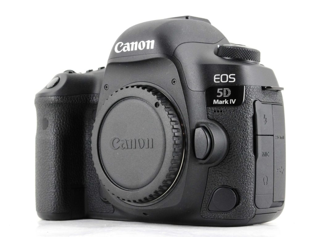 A front and side angled shot of a black Canon EOS 5D Mark IV DSLR camera body with the lens mount cap on. The camera has a textured grip on the right side and the "Canon" and "EOS 5D Mark IV" logos are clearly visible. The background is a solid white.