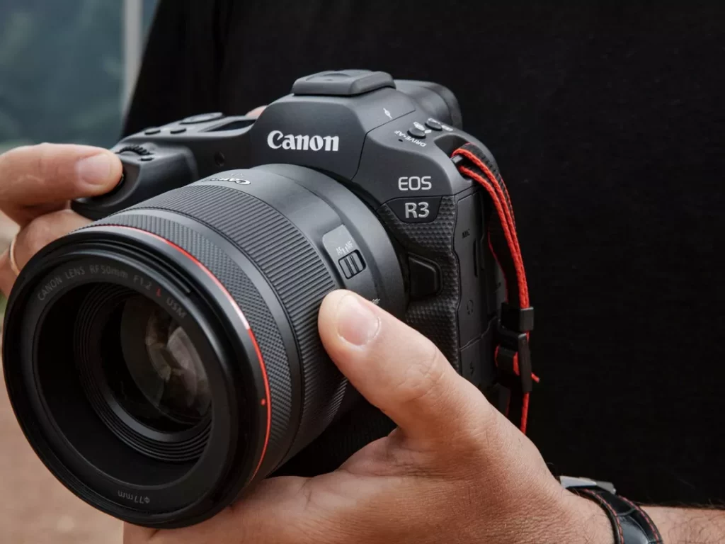 A man holding a professional black Canon EOS R3 mirrorless camera with an RF 50mm F1.2 L USM lens attached. The camera has a red neck strap visible on the right side. The man's hands are adjusting the settings on the camera lens.