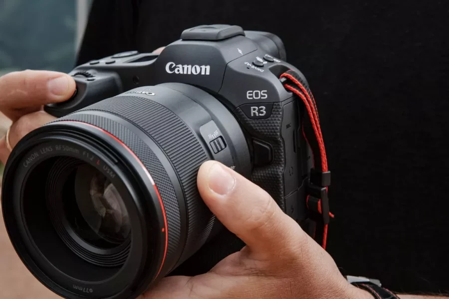 A man holding a professional black Canon EOS R3 mirrorless camera with an RF 50mm F1.2 L USM lens attached. The camera has a red neck strap visible on the right side. The man's hands are adjusting the settings on the camera lens.