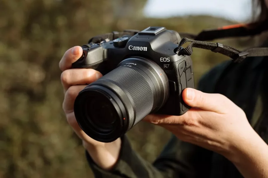 A photographer holds a black Canon EOS R7 mirrorless camera with an RF-S 18-150mm lens attached. The camera is held up and ready to take a picture, with a neck strap visible over the photographer's shoulder. The background is a blurry outdoor scene with trees and foliage.