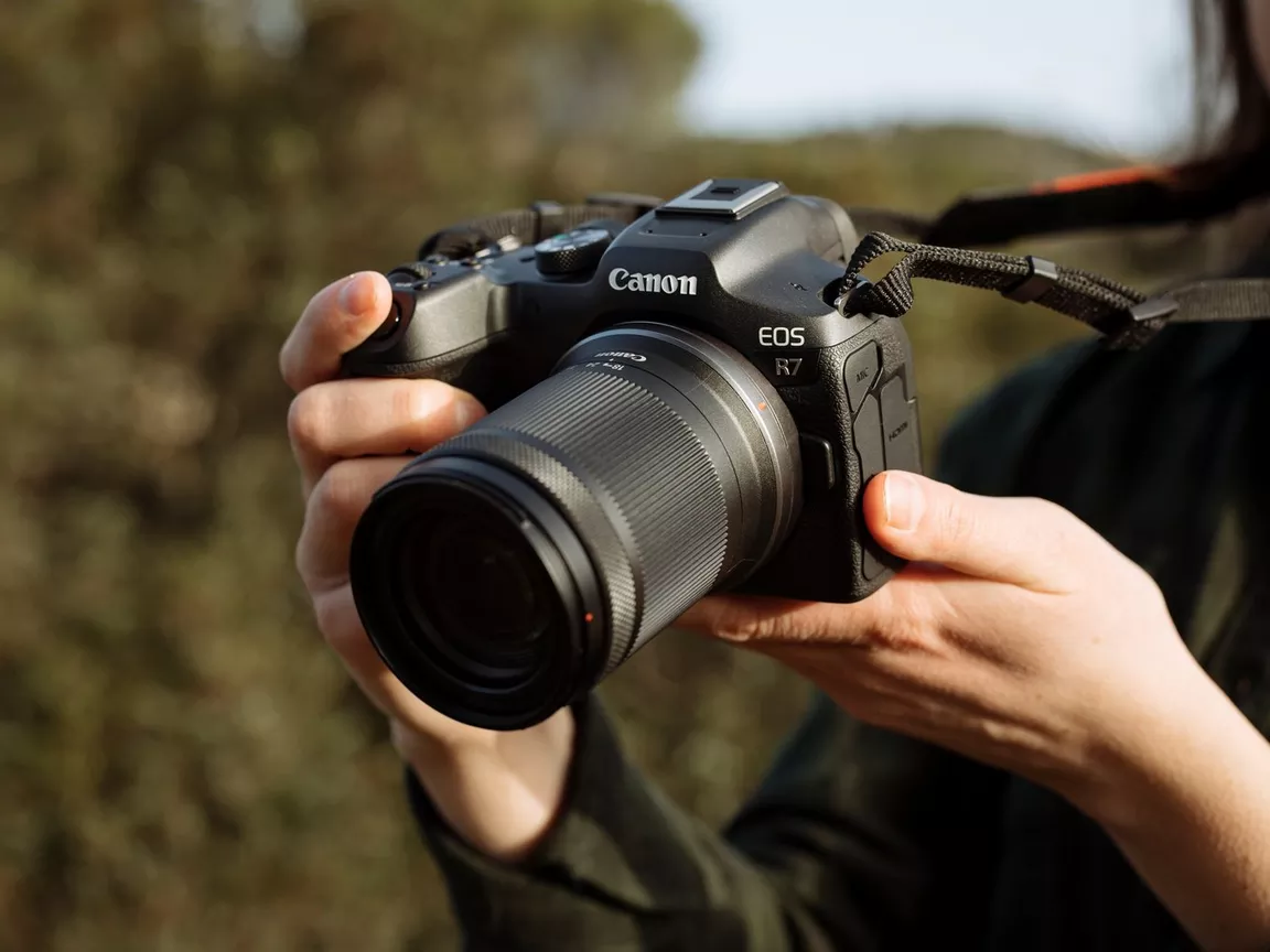 A photographer holds a black Canon EOS R7 mirrorless camera with an RF-S 18-150mm lens attached. The camera is held up and ready to take a picture, with a neck strap visible over the photographer's shoulder. The background is a blurry outdoor scene with trees and foliage.