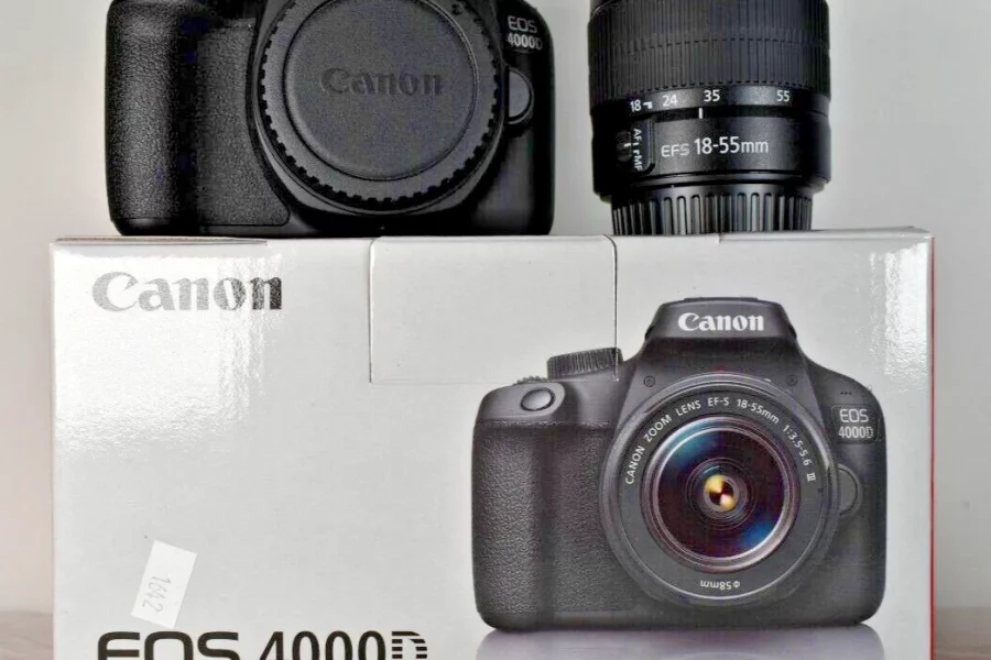 A black Canon EOS 4000D camera body and a Canon EF-S 18-55mm lens sit on top of a white box for the "EOS 4000D EF-S 18-55 III Kit." The camera body has a lens cap on and the lens is standing upright next to it. The box features an image of the camera with the lens attached and has "Canon" and "EOS 4000D" branding, along with some key specifications.
